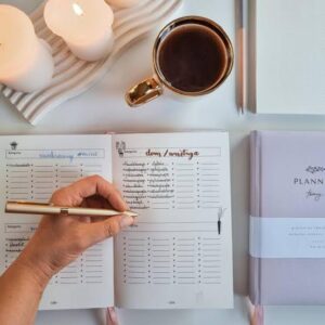 Mulher escrevendo em planner com caneta dourada, acompanhada de xícara de café e velas, representando organização com propósito.