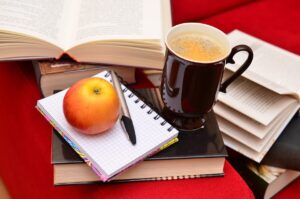 Caderno de anotações, livros empilhados, uma agenda e uma maçã vermelha sobre mesa de madeira com xícara de café. Representando disciplina para o sucesso.